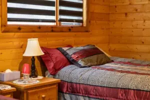 A cozy bedroom in Kitimat features wooden walls and a neatly made bed with a patterned quilt in red, black, and gray tones. A wooden nightstand with a lamp, tissues, and a red cup sits beside the bed. Two windows with blinds provide natural light, perfect for planning your next salmon charter adventure. Skeena Adventure Fishing